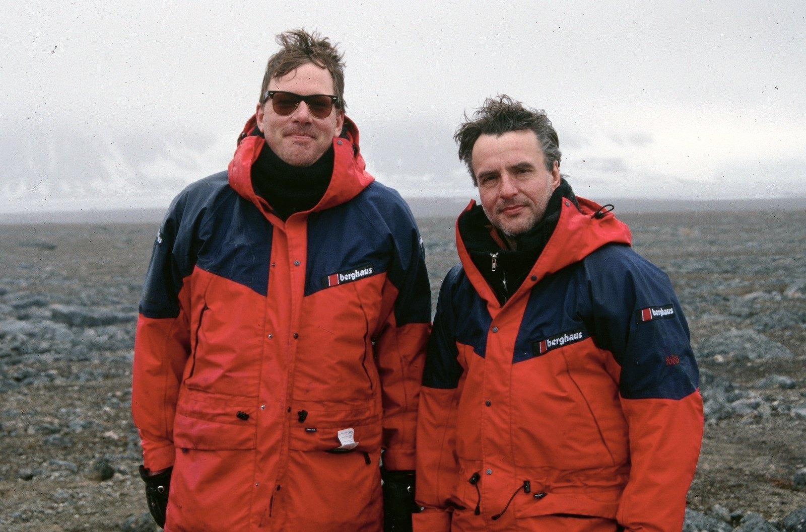 Two men in red and blue parkas standing side by side on a stony shore