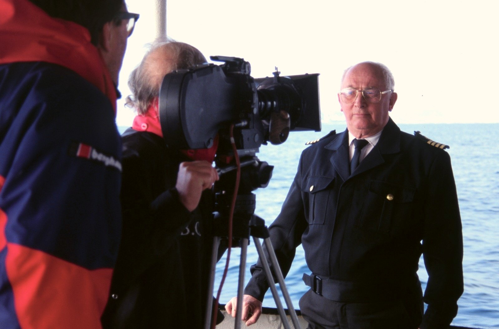 A cameraman filming an officer in a dark uniform on the deck of a ship, the sea behind