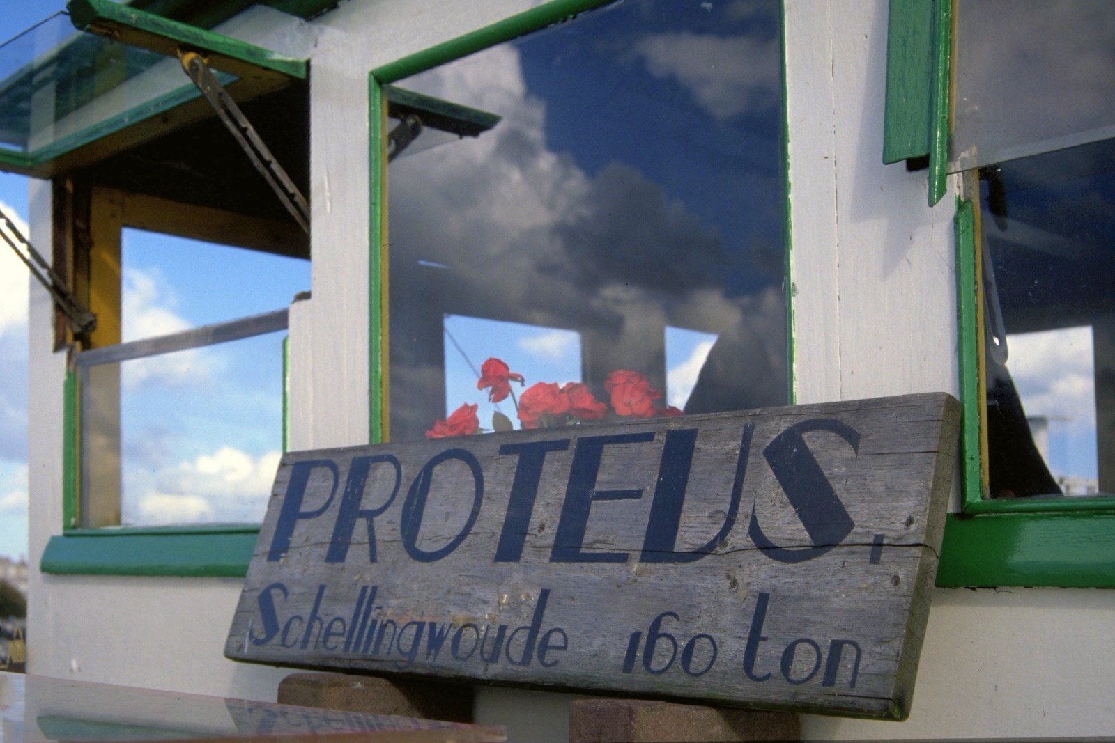 A wooden board painted “PROTEUS Schellingwoude 160 ton” on a boat’s wheelhouse with red geraniums