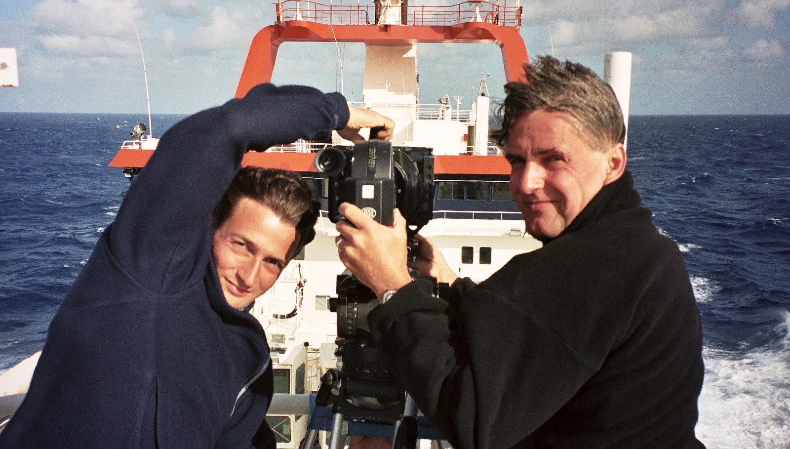 Two men with a film camera on the stern deck of a ship, the open sea and the white superstructure behind