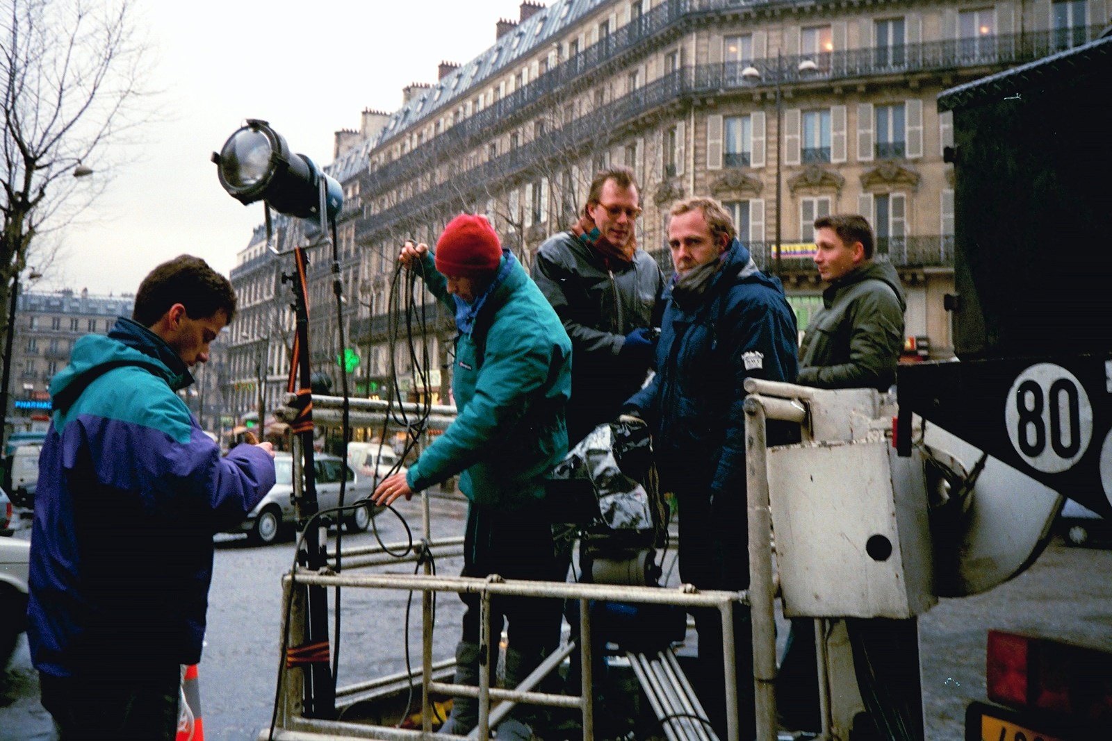 Crew members in winter jackets around a film lamp on a stand in a Paris street, apartment buildings behind