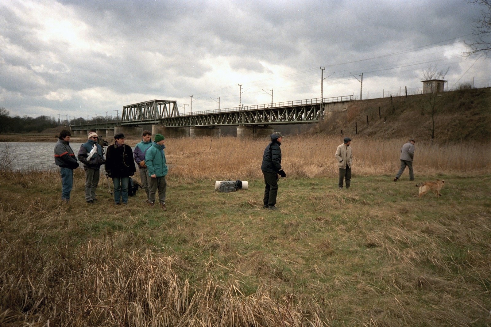 A group of people standing in a field of dry grass, a steel railway bridge on the horizon under heavy clouds