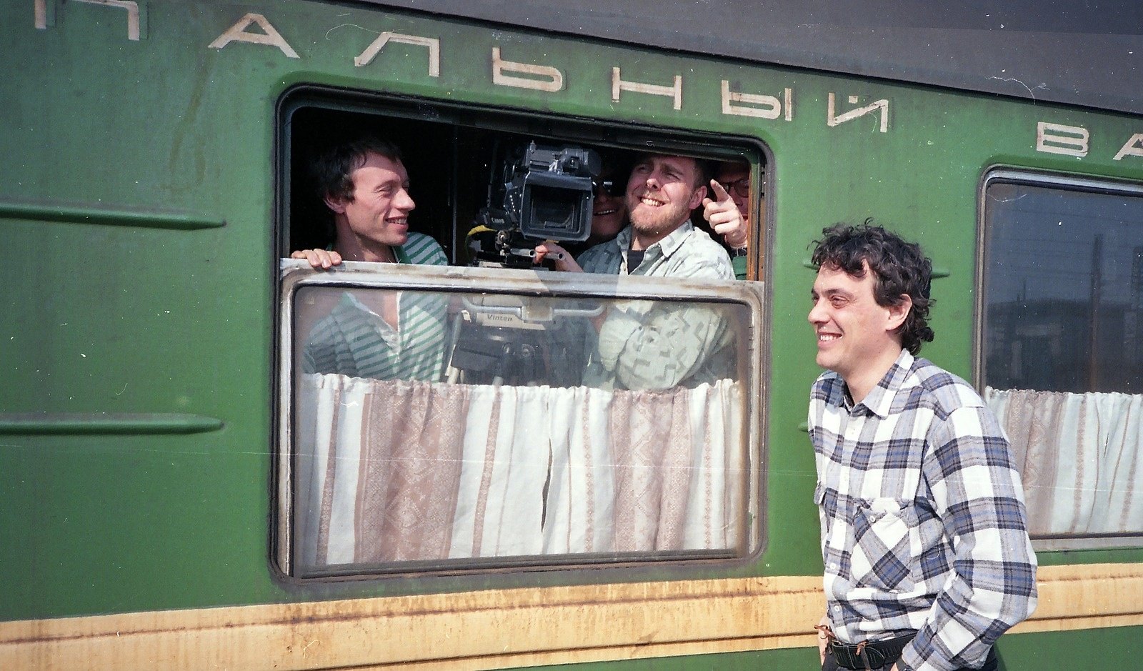 Two men with a film camera leaning out of the window of a green Soviet carriage, a third man standing outside