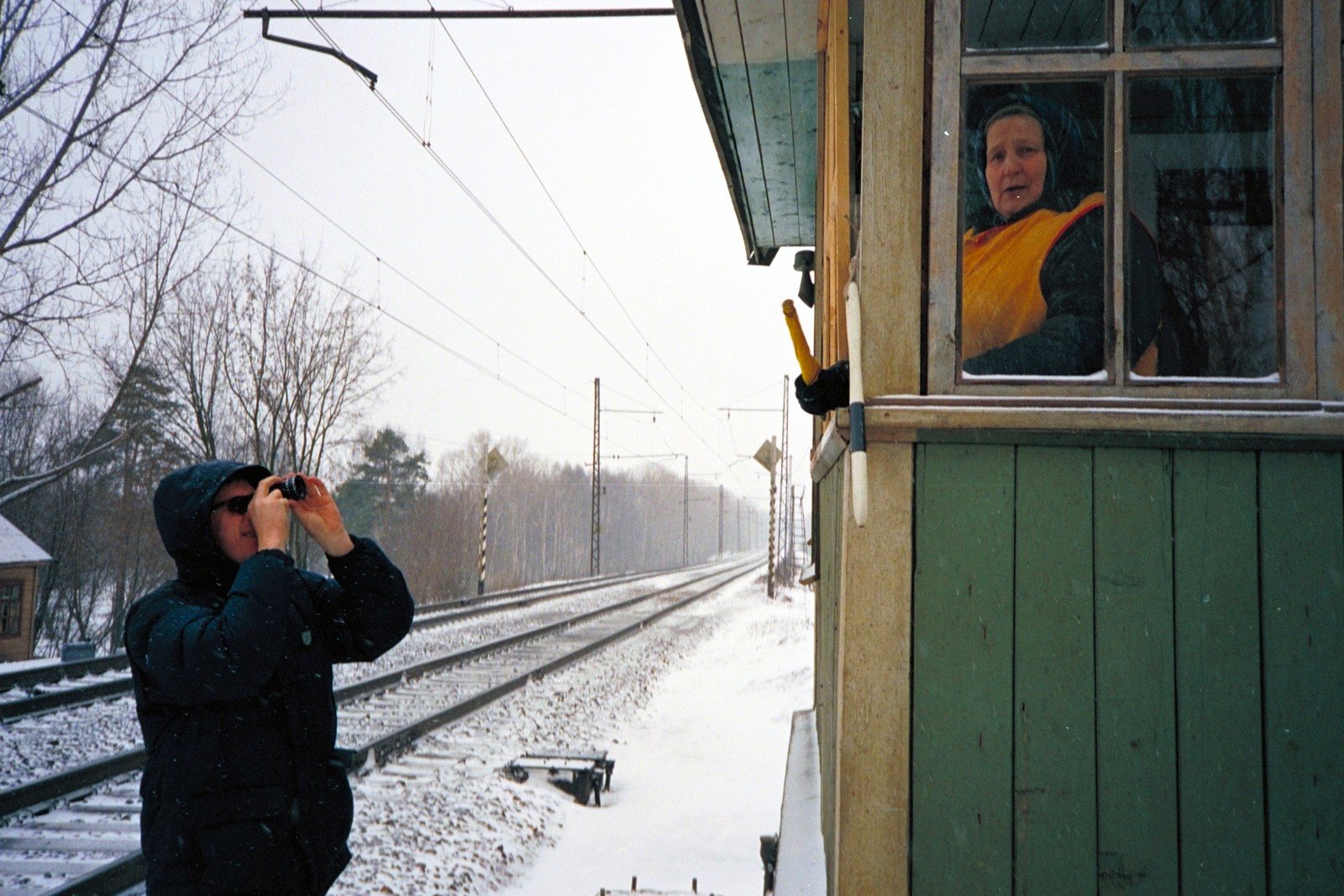 A man in a hooded jacket on snowy railway tracks, looking through a camera; a woman watches from the window of a green signal box