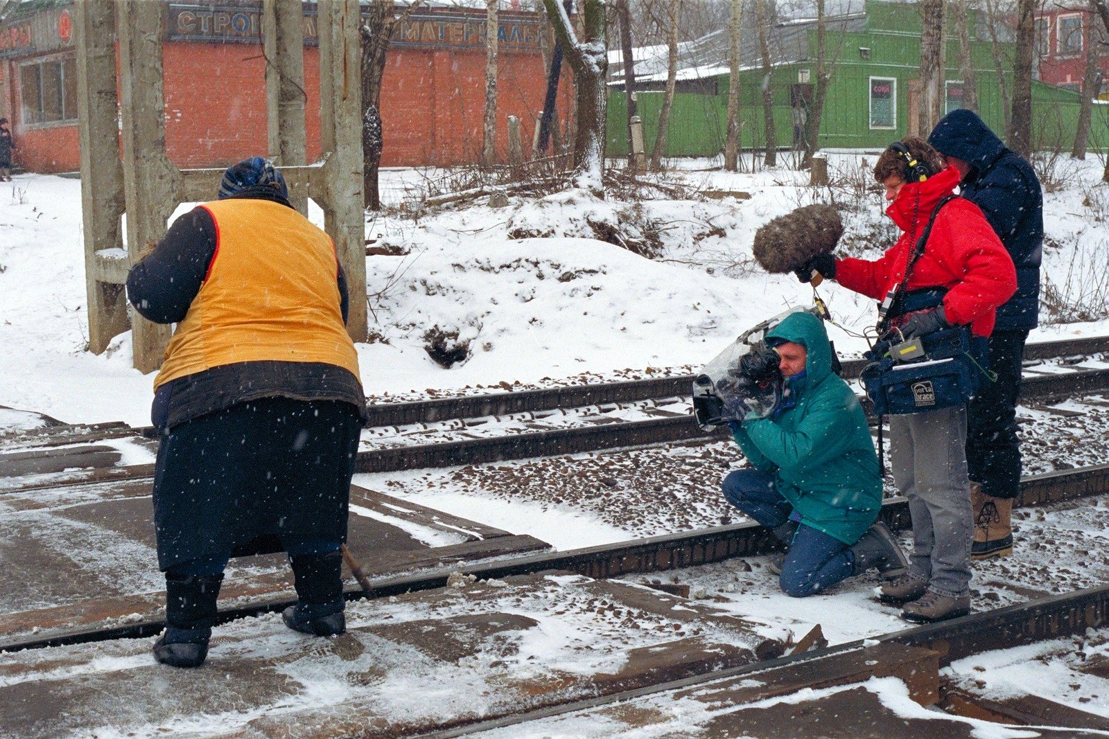 In the snow by railway tracks, a cameraman kneels with the camera while a sound recordist holds a microphone; a woman in an orange vest stands nearby