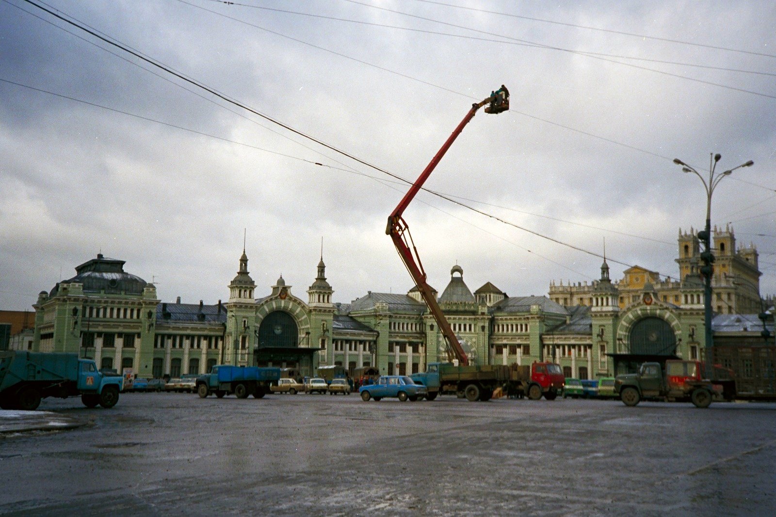 A wet square in front of a long green and white station building with towers, a red crane arm raised above parked lorries