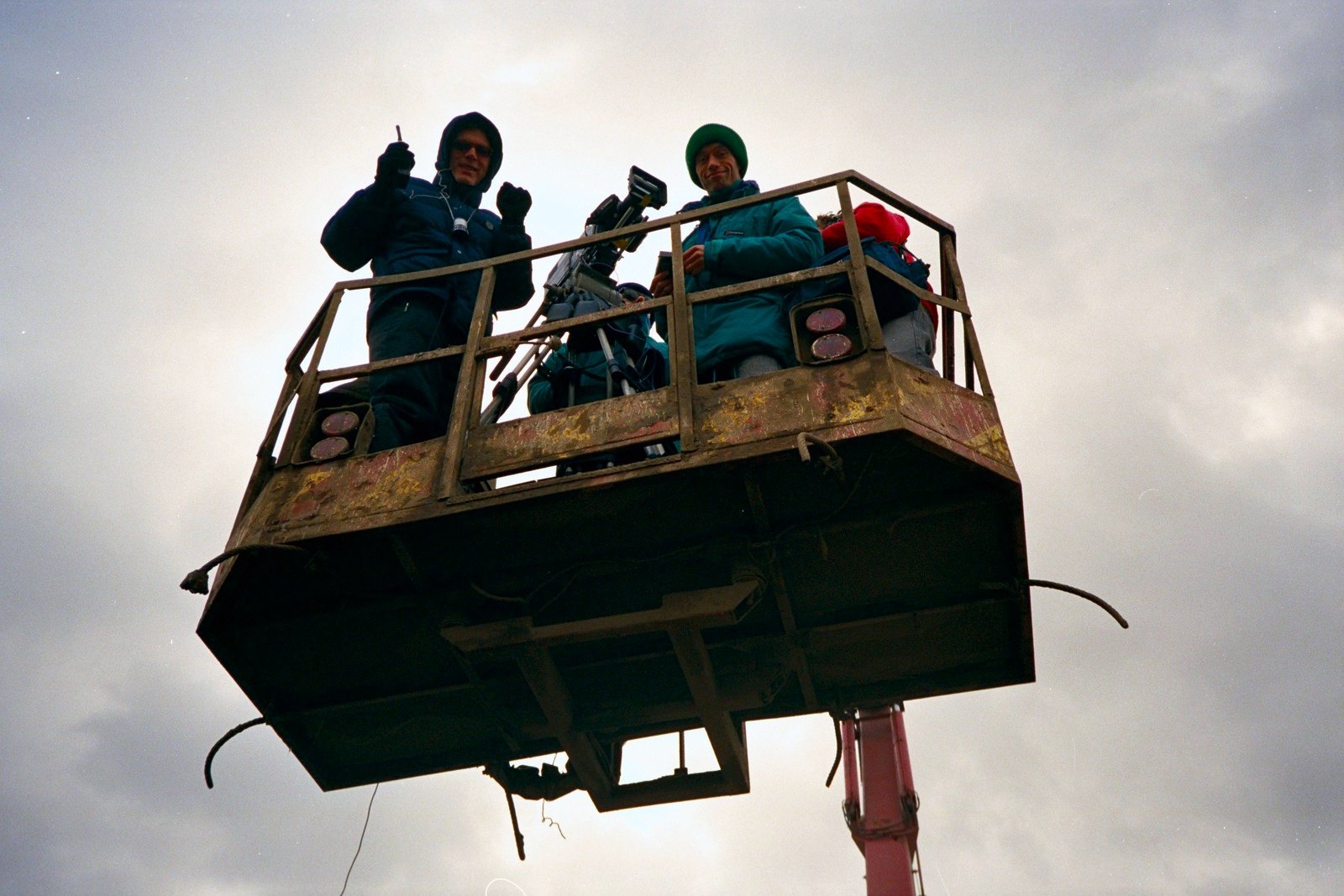 Seen from below, three people with a camera on the platform of a crane, against a pale sky