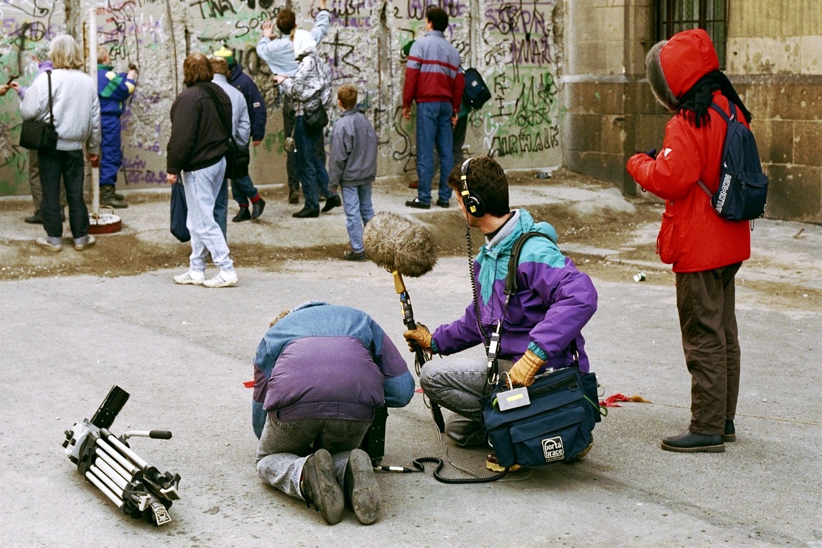 People chipping at a stretch of the Berlin Wall covered in graffiti; in front, a sound recordist with a boom crouches beside a kneeling man, a tripod on the ground