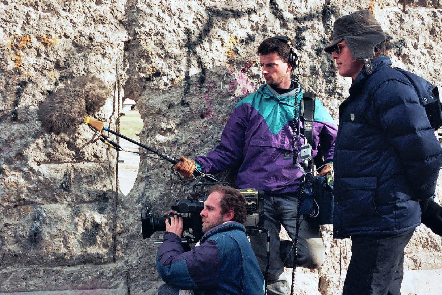 At the Berlin Wall, a cameraman crouching with a camera, a sound recordist with a boom behind him and a man in a cap and dark jacket beside them