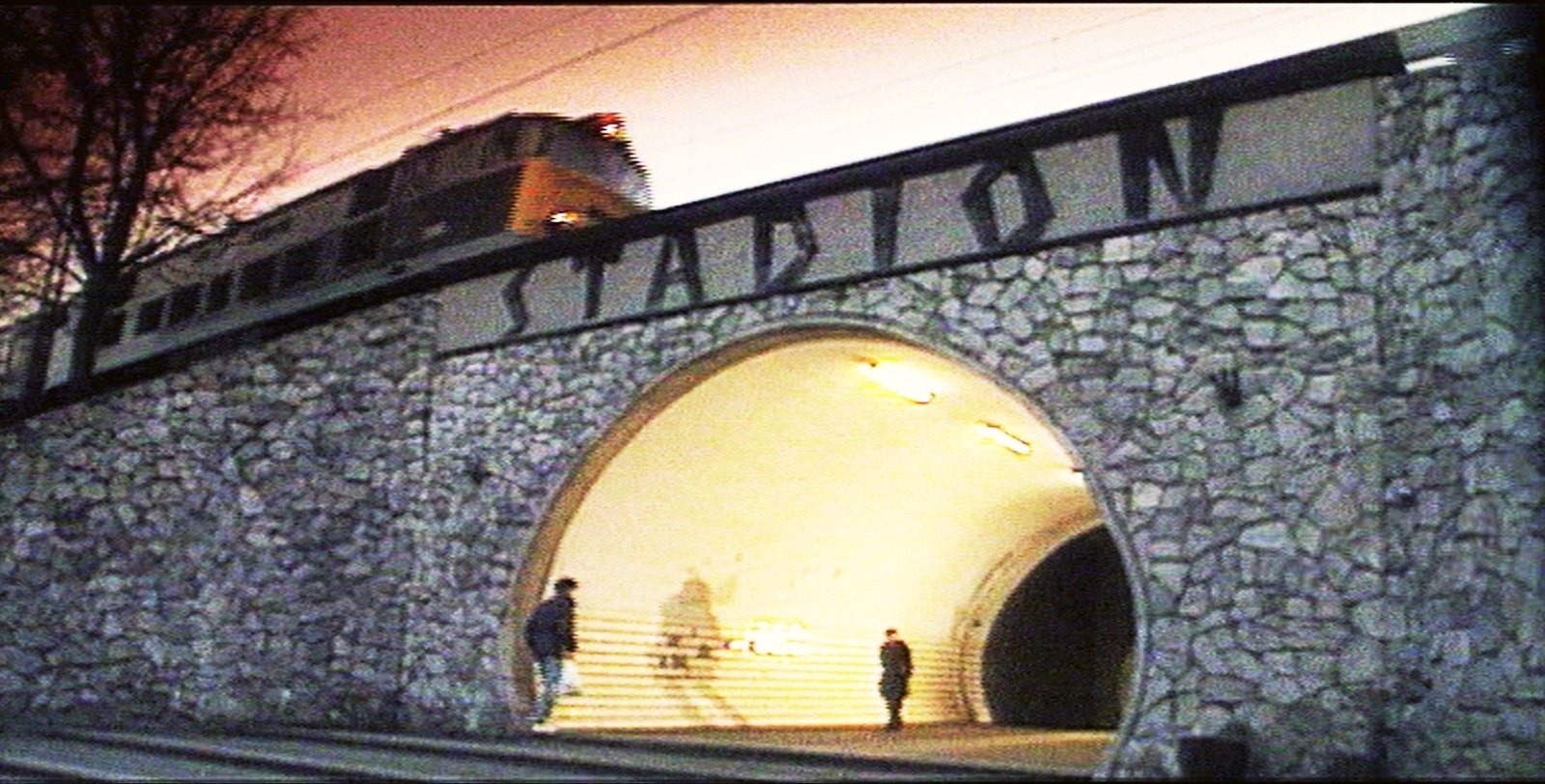 Dusk at a stone railway viaduct with the word STADION in large letters, a train passing above and two figures in the lit arched passage
