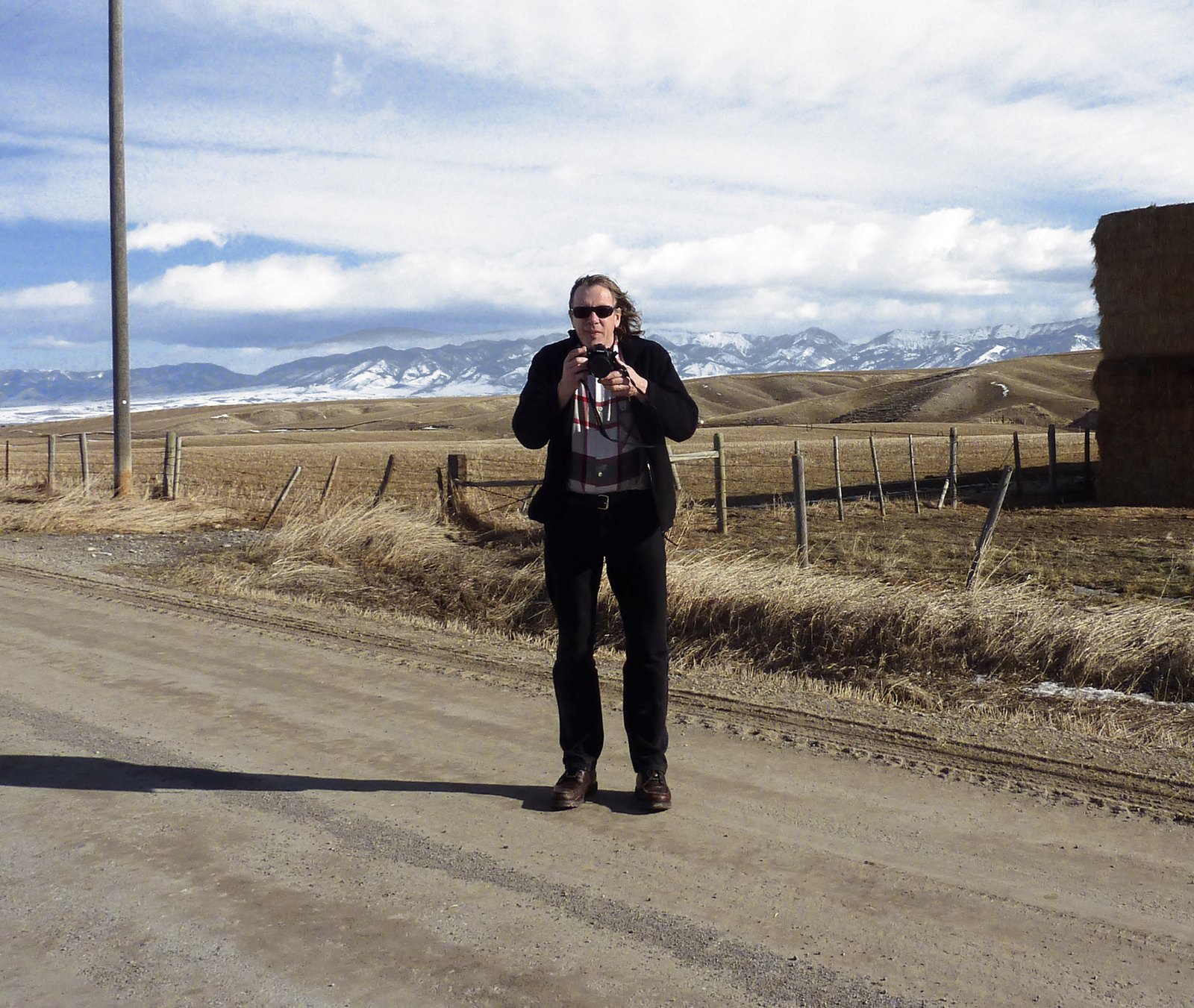 Rob Rombout standing on a gravel road in Montana, camera raised to his eye, with fences, a stack of hay bales and snowy mountains behind him